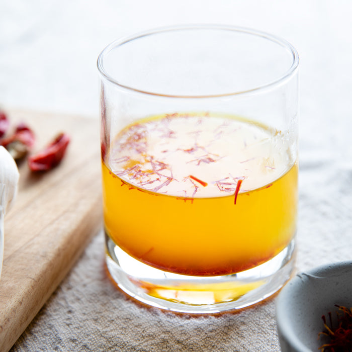 Close-up of glass of saffron-infused oil with saffron threads, cutting board with garlic and dried chiles blurred behind