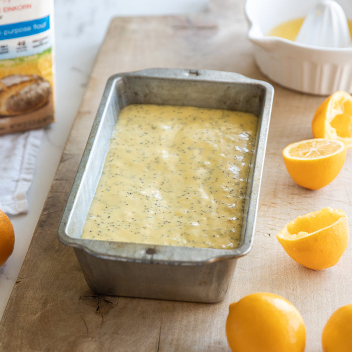 Einkorn lemon poppy seed loaf batter in a metal loaf pan on a wooden board with halved lemons and flour bag