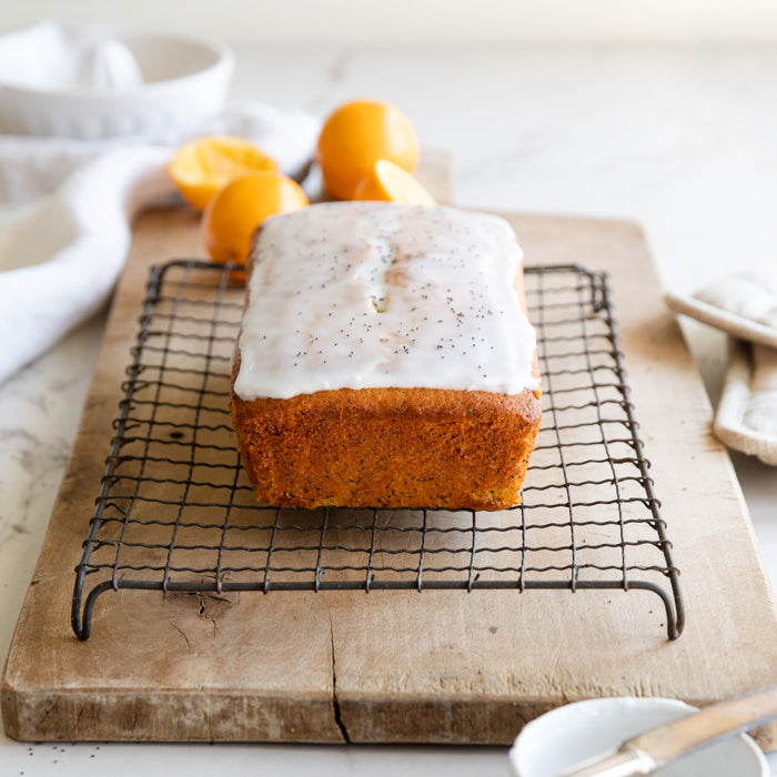 Front view of Einkorn lemon poppy seed loaf with glaze on a cooling rack, halved lemons behind