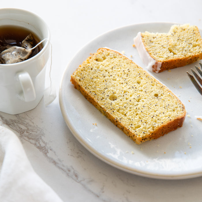 Top-down view of sliced Einkorn lemon poppy seed loaf showing poppy seeds and glaze on a white plate with a mug of tea