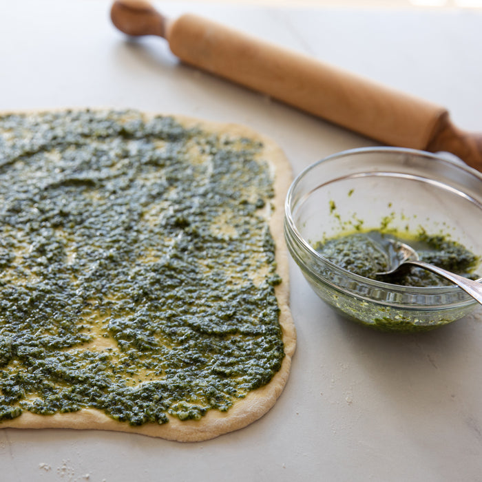 Rolled-out Einkorn pizza dough spread with green pesto, a glass bowl of pesto and a wooden rolling pin on a light countertop