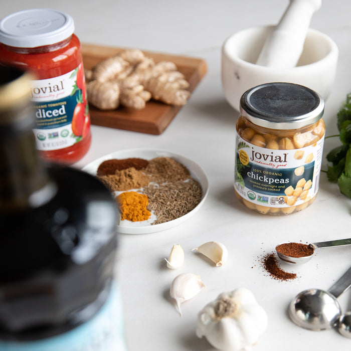 Jovial organic chickpeas jar with spices, garlic, a jar of diced tomatoes, and mortar on a white counter for vegan curry