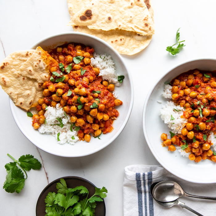 Two bowls of vegan chickpea curry over rice with torn flatbread and fresh cilantro on a white surface