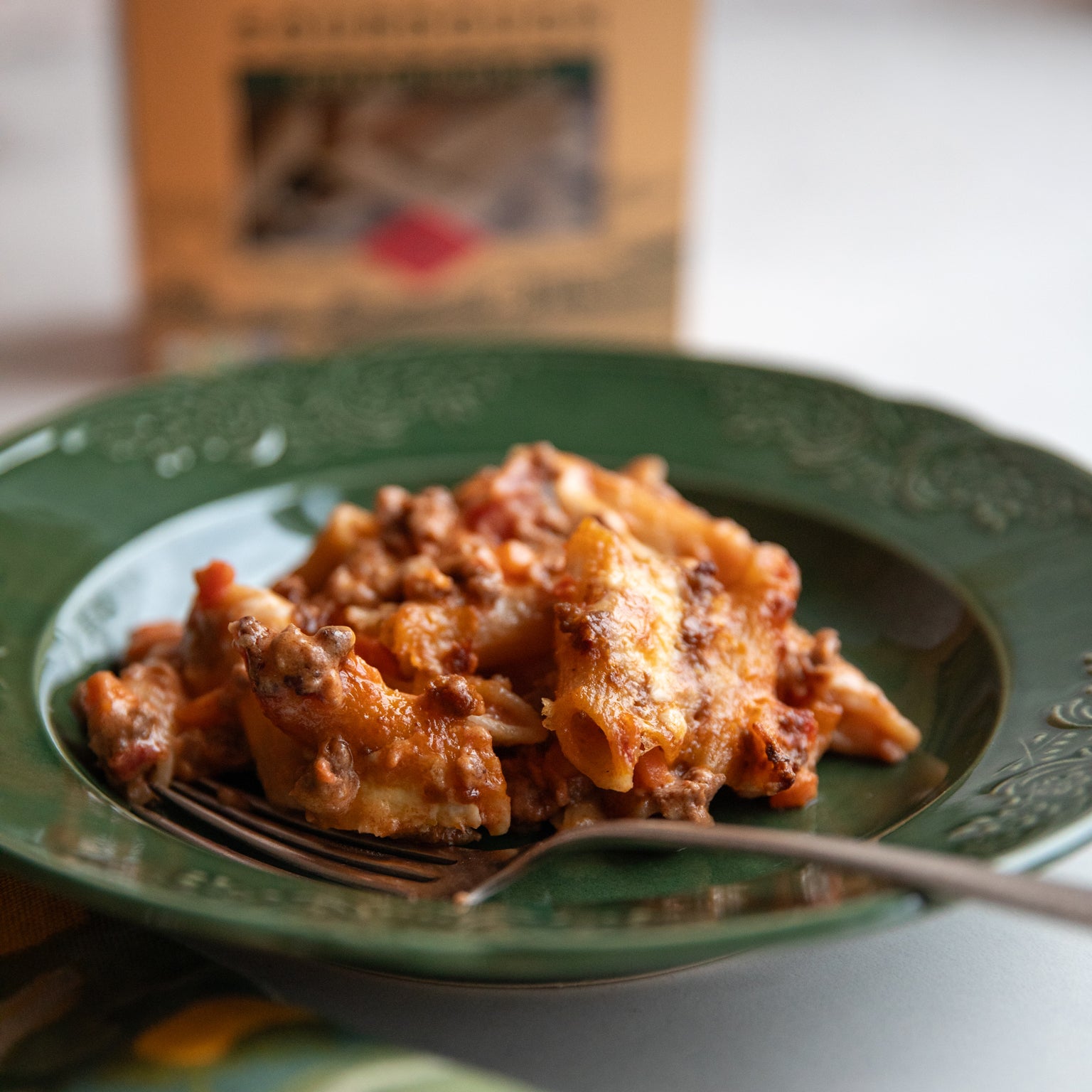 Sourdough penne al forno on a green plate with browned cheese and meat sauce, fork and blurred pasta box in background