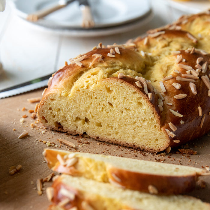 Close-up of sliced Einkorn Easter bread showing tender yellow crumb, golden crust, and slivered almonds on parchment with blurred dishes behind