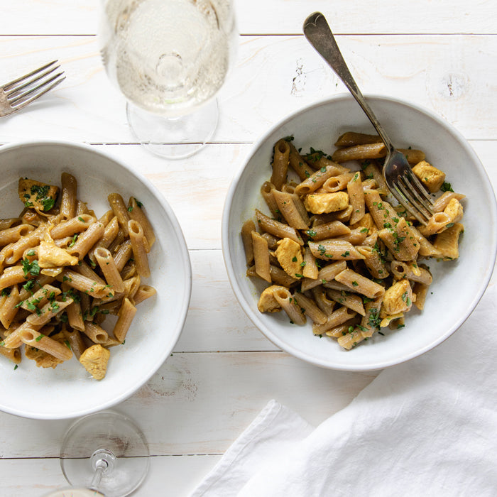 Two bowls of organic cassava penne with coconut-curried chicken and herbs on a white cloth with forks and a wine glass