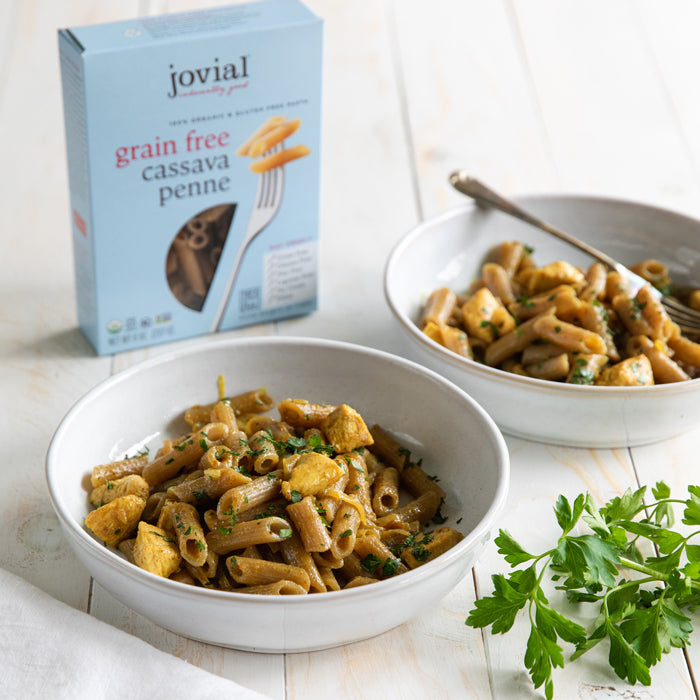 Two bowls of coconut curry chicken with Jovial cassava penne on a light wood table with Jovial box and parsley garnish