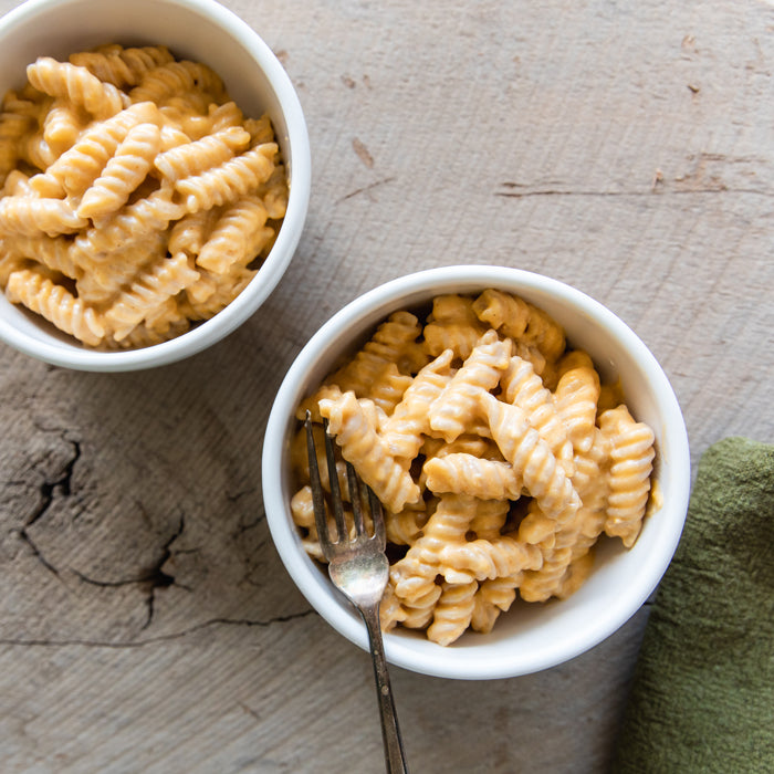 Two bowls of creamy fusilli mac and cheese on a wooden surface, fork in the nearest bowl and a green napkin beside it