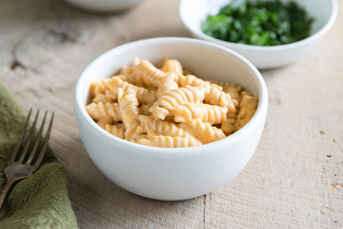 Close-up of fusilli in creamy cheese sauce in a white bowl with green napkin and blurred herbs behind