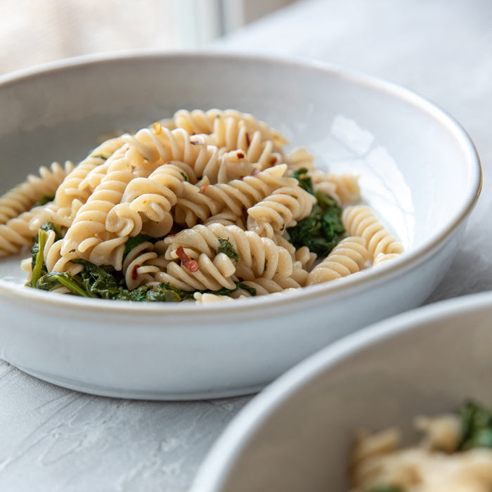 Close-up of fusilli pasta with spring greens in a white bowl, showing twisted spirals and wilted greens on a light tabletop