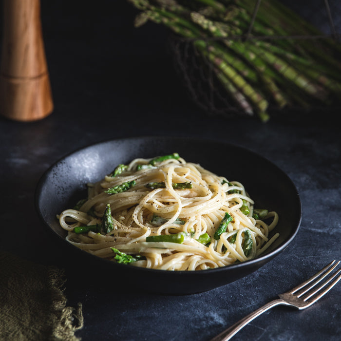 Black bowl of linguine tossed with asparagus, creamy cheese, and cracked black pepper on a dark tabletop