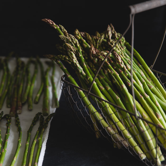 Close-up of fresh asparagus spears in a wire basket with extra spears on a white cloth and dark background