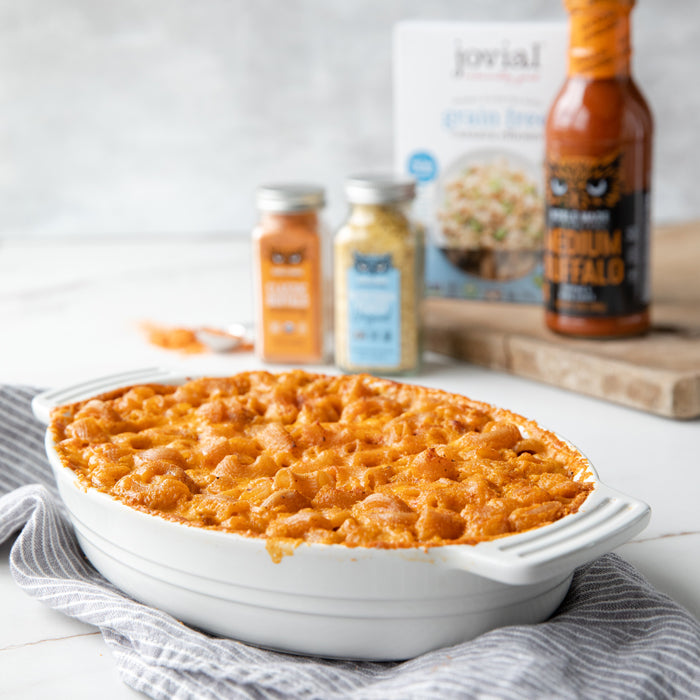 Buffalo mac and cheese in a white oval baking dish on a striped towel, Jovial product boxes and hot sauce blurred behind