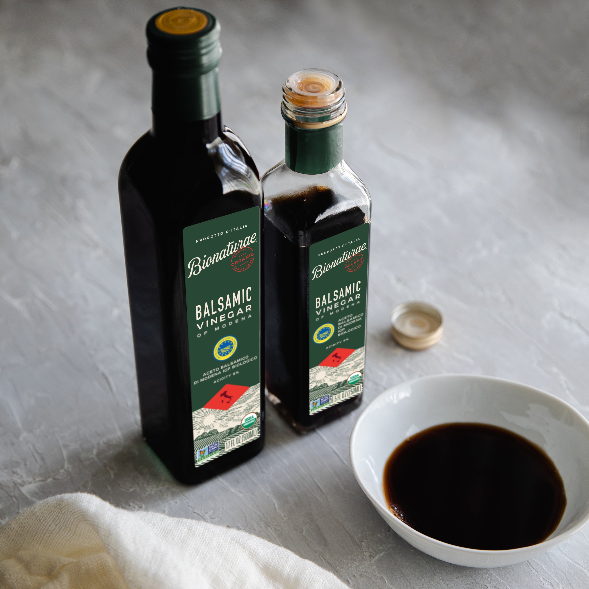 Three-quarter view of Bionaturae organic balsamic vinegar 17 fl oz bottles showing green labels and certification badges with a white dipping bowl on a gray tabletop