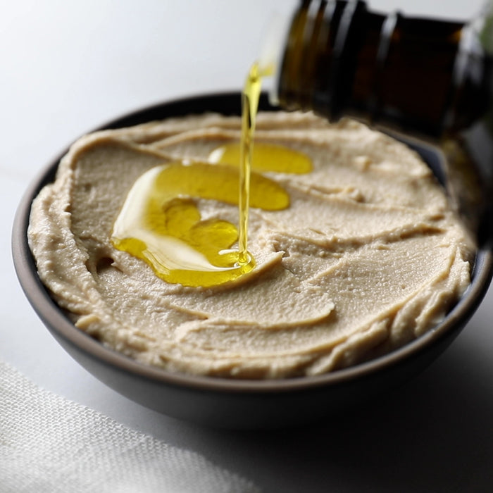 Close-up top view of hummus in a dark bowl showcasing smooth, whipped texture and olive oil drizzle being poured with soft white background
