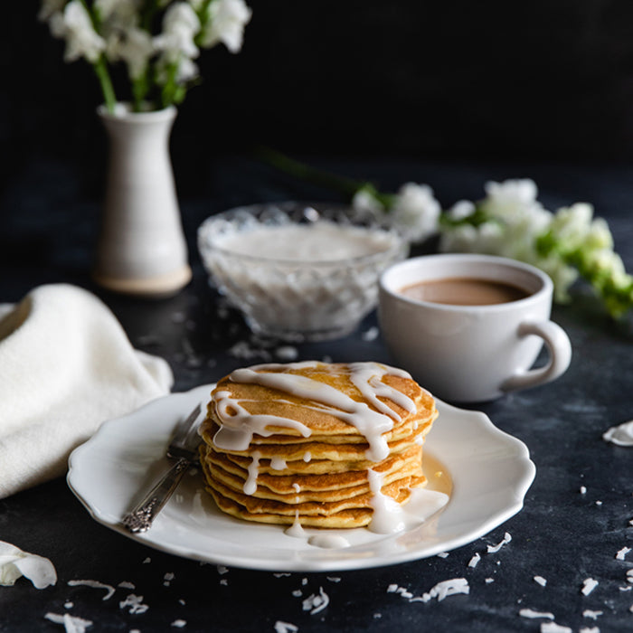Stack of dairy-free Einkorn coconut pancakes drizzled with coconut sauce on a white plate with a cup of coffee and flowers