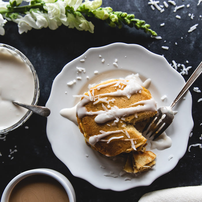 Dairy-free einkorn coconut pancakes on a white plate drizzled with coconut sauce and shredded coconut, fork and coffee nearby