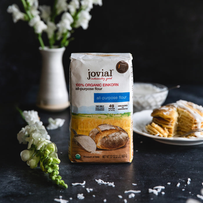 Stack of dairy-free einkorn coconut pancakes topped with shredded coconut and sauce, plated beside a bag of Jovial organic einkorn flour on a dark tabletop