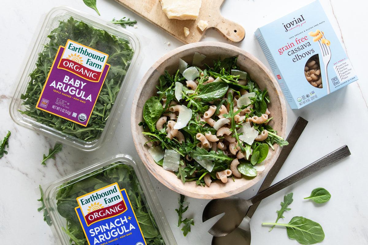 Pasta salad with arugula and shaved Parmesan in wooden bowl with Earthbound Farm arugula and Jovial cassava elbows box