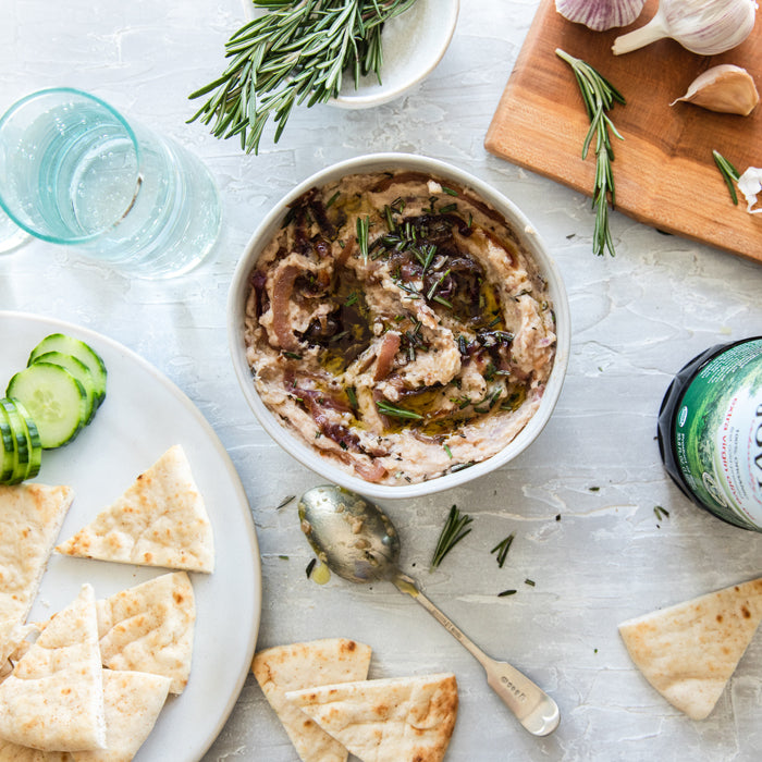 Bowl of caramelized onion white bean dip drizzled with olive oil and rosemary, with pita wedges and cucumber slices