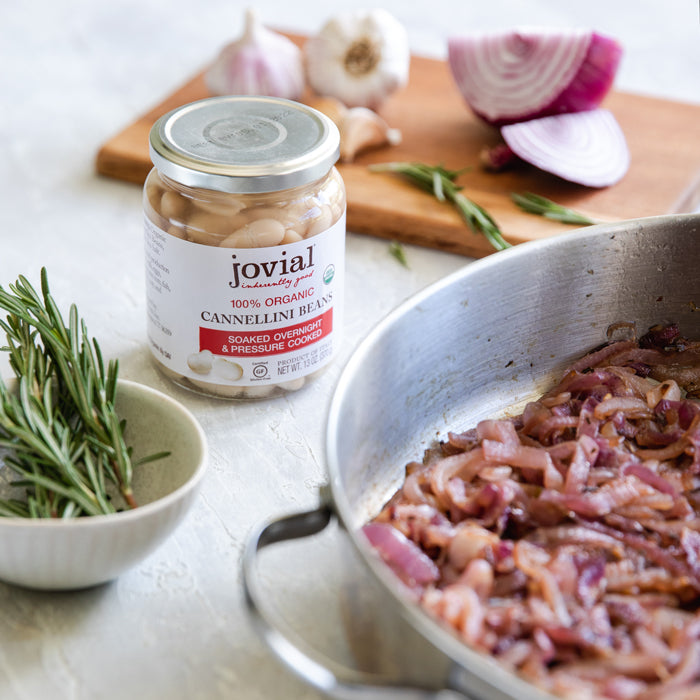Jar of Jovial organic cannellini beans beside a skillet of caramelized red onions with rosemary and garlic on a kitchen counter