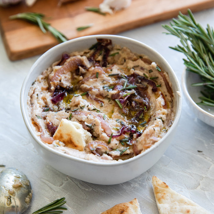 Overhead view of caramelized onion white bean dip in a bowl topped with rosemary and olive oil, pita pieces at side