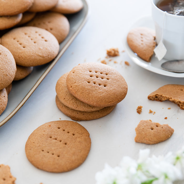 Stacked homemade Einkorn digestive biscuits beside a cup of tea on a white surface with scattered crumbs