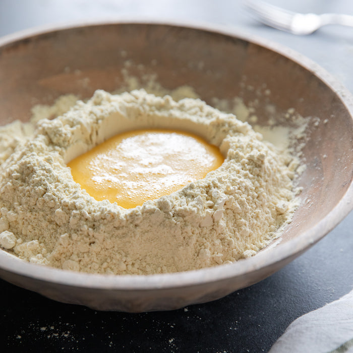 Bowl of einkorn flour with eggs in a well ready to be mixed into fresh pasta dough on a dark countertop