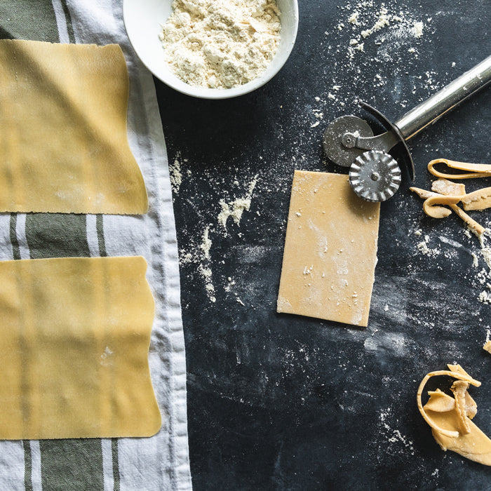 Einkorn pasta dough rolled into thin sheets on a flour-dusted black counter with a pastry wheel and bowl of flour