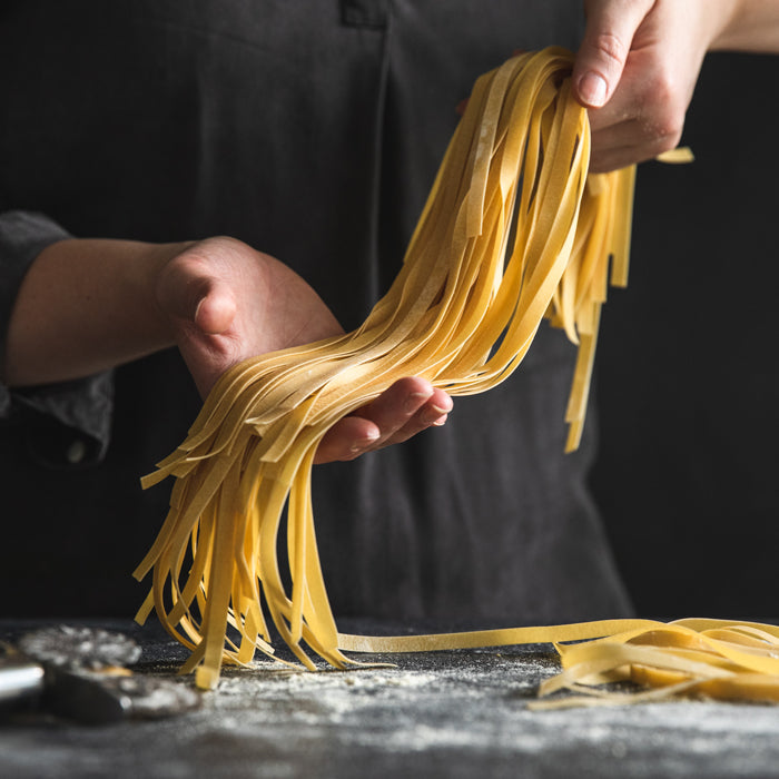 Hands lifting fresh einkorn ribbon pasta over a floured countertop against a dark background