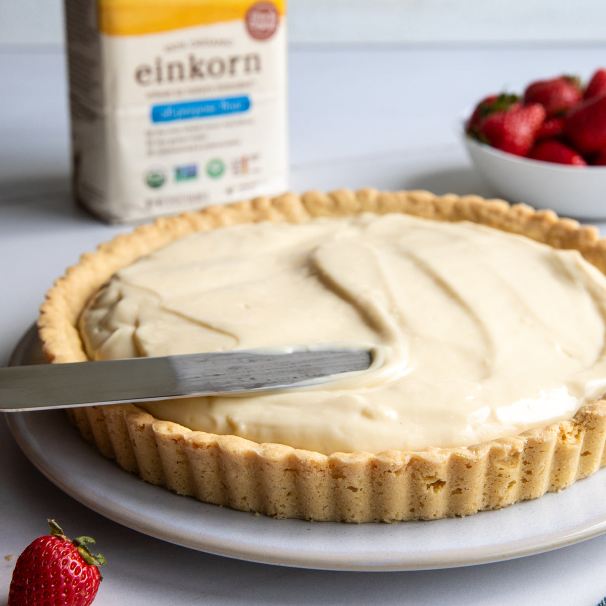 Close-up of 9-inch Einkorn tart with pastry cream in a fluted crust, bowl of strawberries and flour bag behind