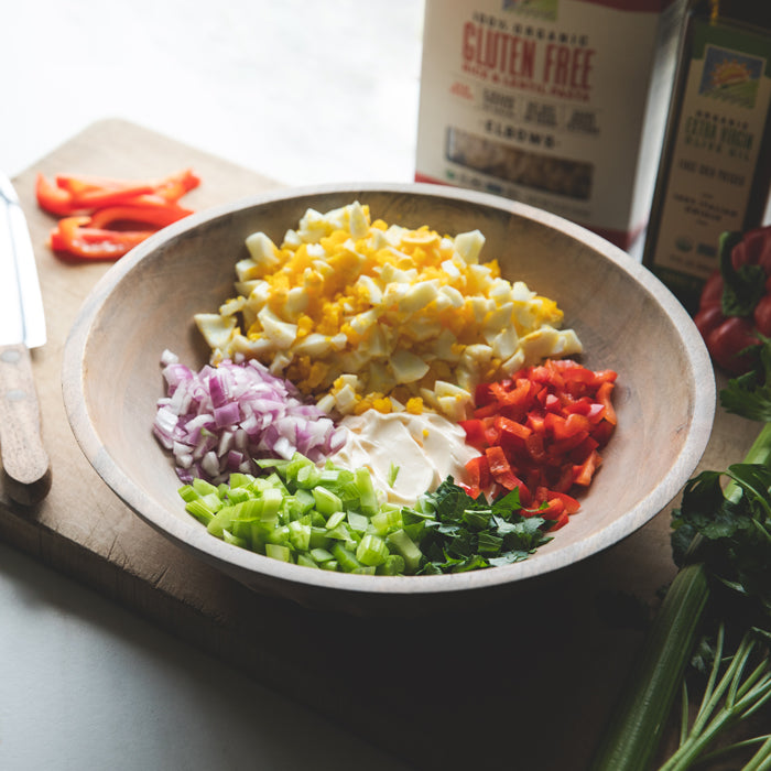 Macaroni salad ingredients in wooden bowl: chopped egg, red pepper, celery and mayo, gluten-free pasta box in background