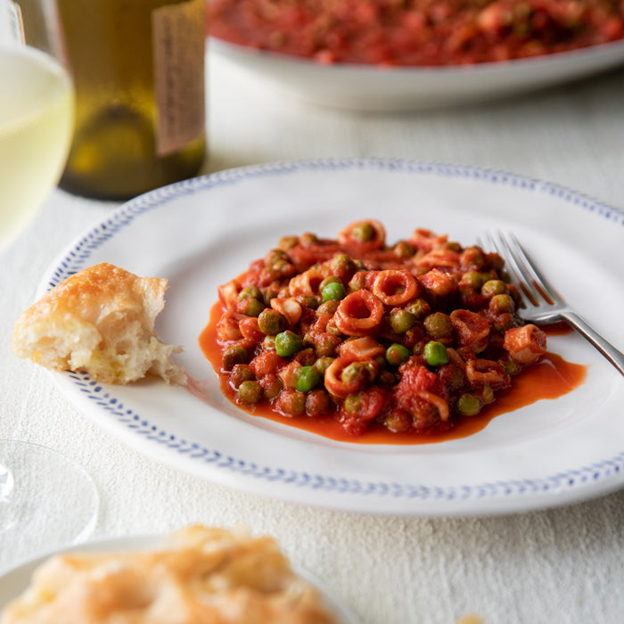 Calamari with peas in tomato sauce on a white plate with torn bread and a glass of white wine in the background