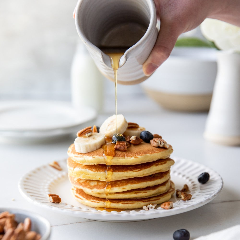 Stack of golden organic Einkorn pancakes topped with butter and blueberries as syrup is poured over them on a white plate