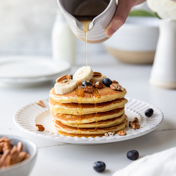Stack of golden einkorn pancakes with banana slices, pecans, and blueberries as syrup is poured over them on a white plate