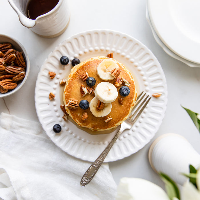 Stack of golden einkorn pancakes topped with banana slices, blueberries, and chopped pecans on a white plate