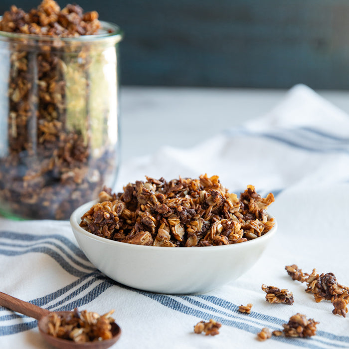 Front view of chocolate Einkorn wheat berry granola in a white bowl showing crunchy clusters on a blue-striped cloth