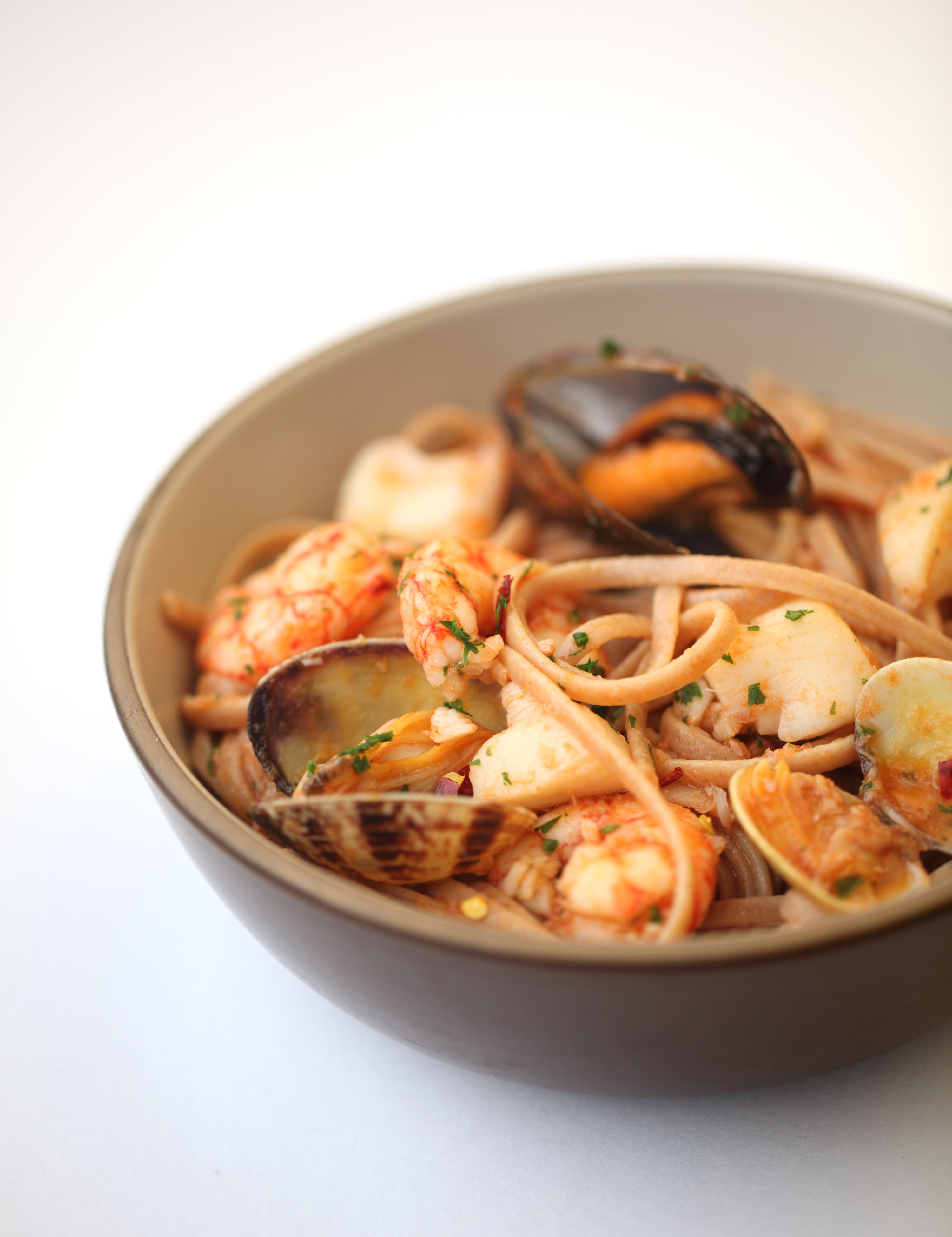 Close-up of einkorn linguine with mussels, clams, and shrimp in tomato broth in a brown bowl on a white background