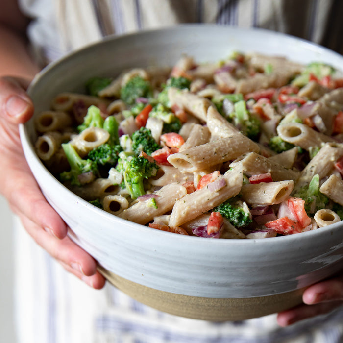 Gluten-free hummus pasta salad in a ceramic bowl held by a person, with broccoli, red pepper, and red onion