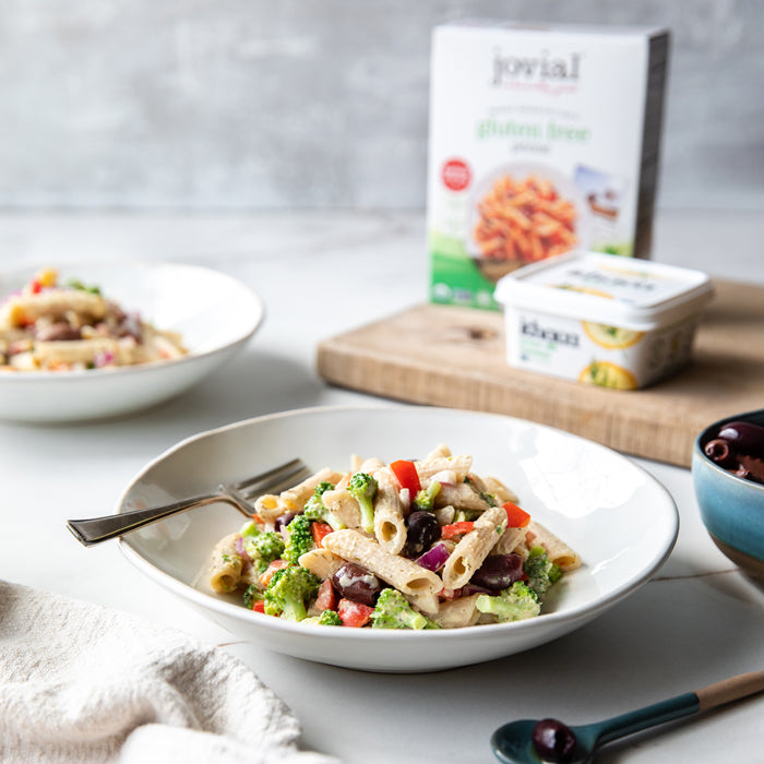 Gluten-free hummus pasta salad with penne, broccoli, red pepper and olives, Jovial pasta box blurred in background