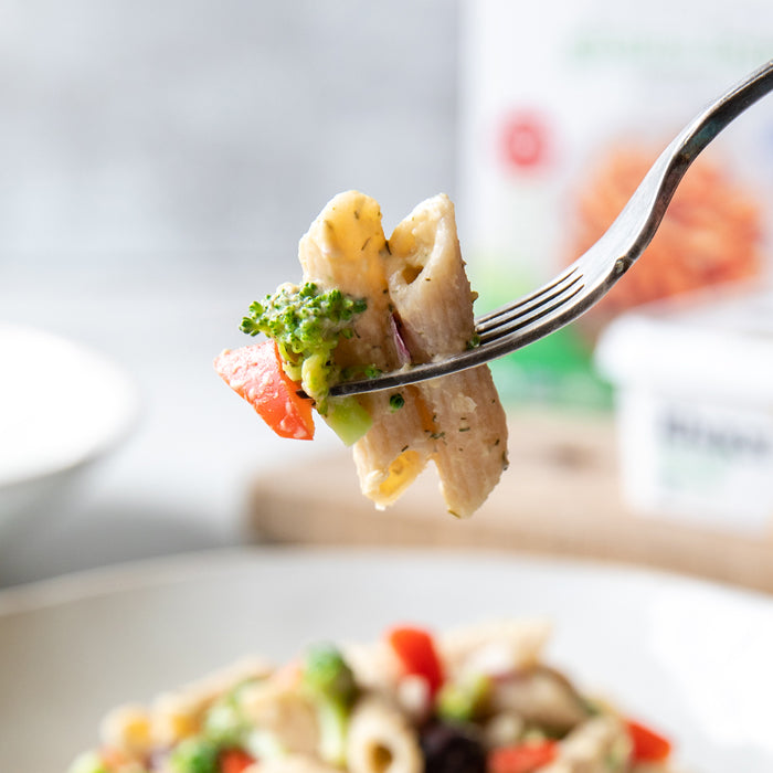 Close up of a fork lifting a bite of gluten free hummus based pasta salad with broccoli and red pepper over a blurred bowl