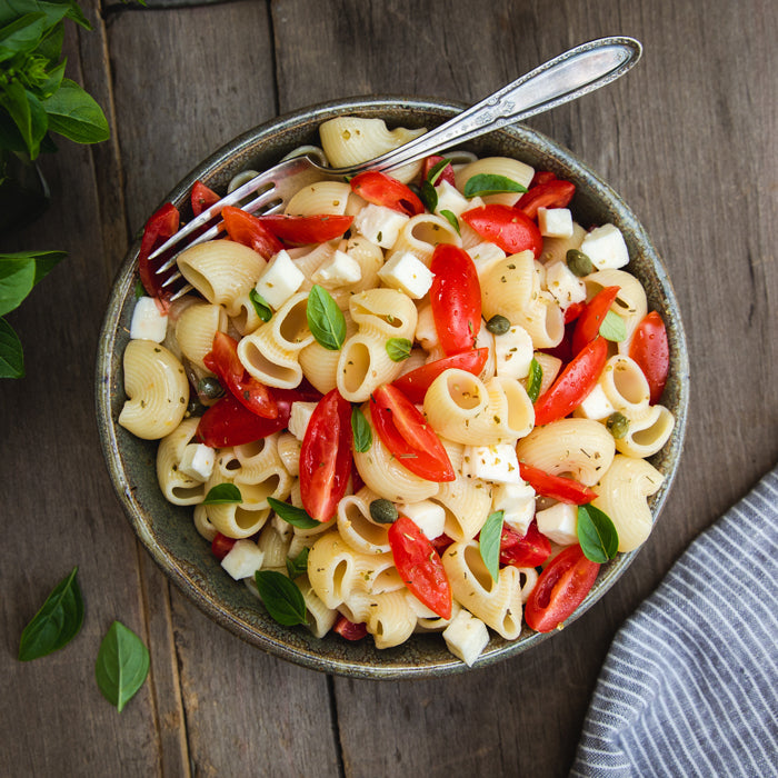 Bowl of chiocciole pasta with cherry tomatoes, cubed mozzarella, basil leaves and capers on a wooden table