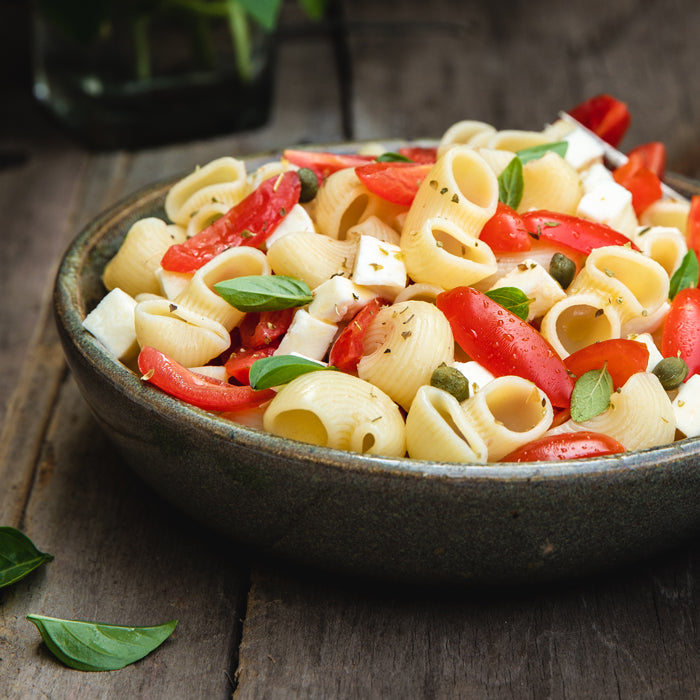 Chiocciole pasta salad in a ceramic bowl with cherry tomatoes, diced mozzarella, basil leaves, and capers on a rustic wooden table