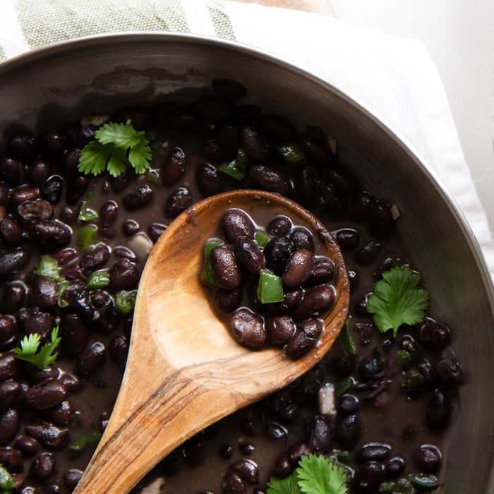 Close-up of a wooden spoon scooping cooked black beans with diced jalapeño and cilantro from a dark pot on a white cloth