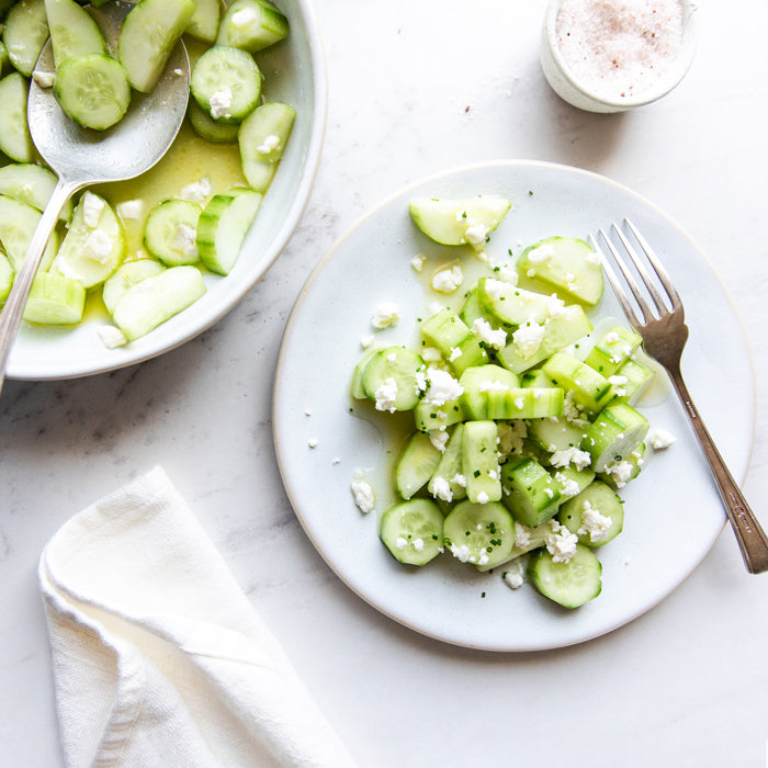Fresh cucumber and goat cheese salad served on a white plate with a bowl of extra salad and a fork on a marble surface