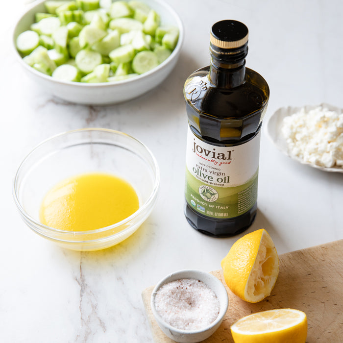 Ingredients for cucumber goat cheese salad on a marble countertop: sliced cucumbers, bottle of Jovial extra virgin olive oil, lemon, goat cheese, salt, and dressing