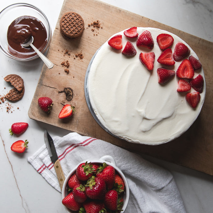 Top-down view of ice cream cake topped with halved strawberries, with a bowl of strawberries, chocolate sauce, and Einkorn crispy cocoa cookies on a wooden board
