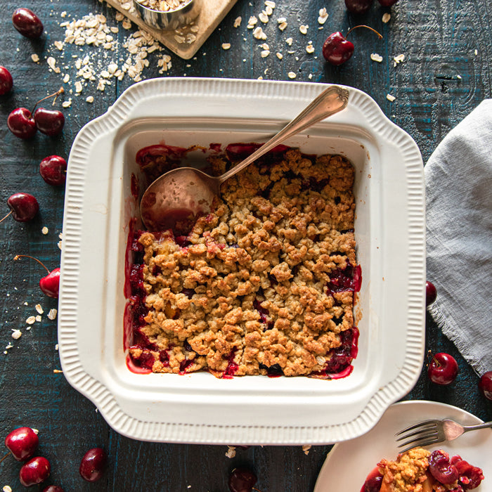 Einkorn fruit crumble in a white baking dish with a serving spoon, fresh cherries and oats scattered on dark wooden table