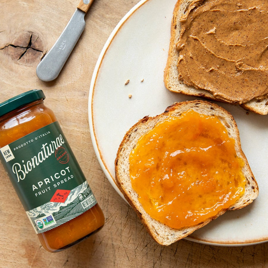Overhead view of Bionaturae Organic Apricot Fruit Spread jar showcasing a slice of toast topped with apricot spread on a wooden board