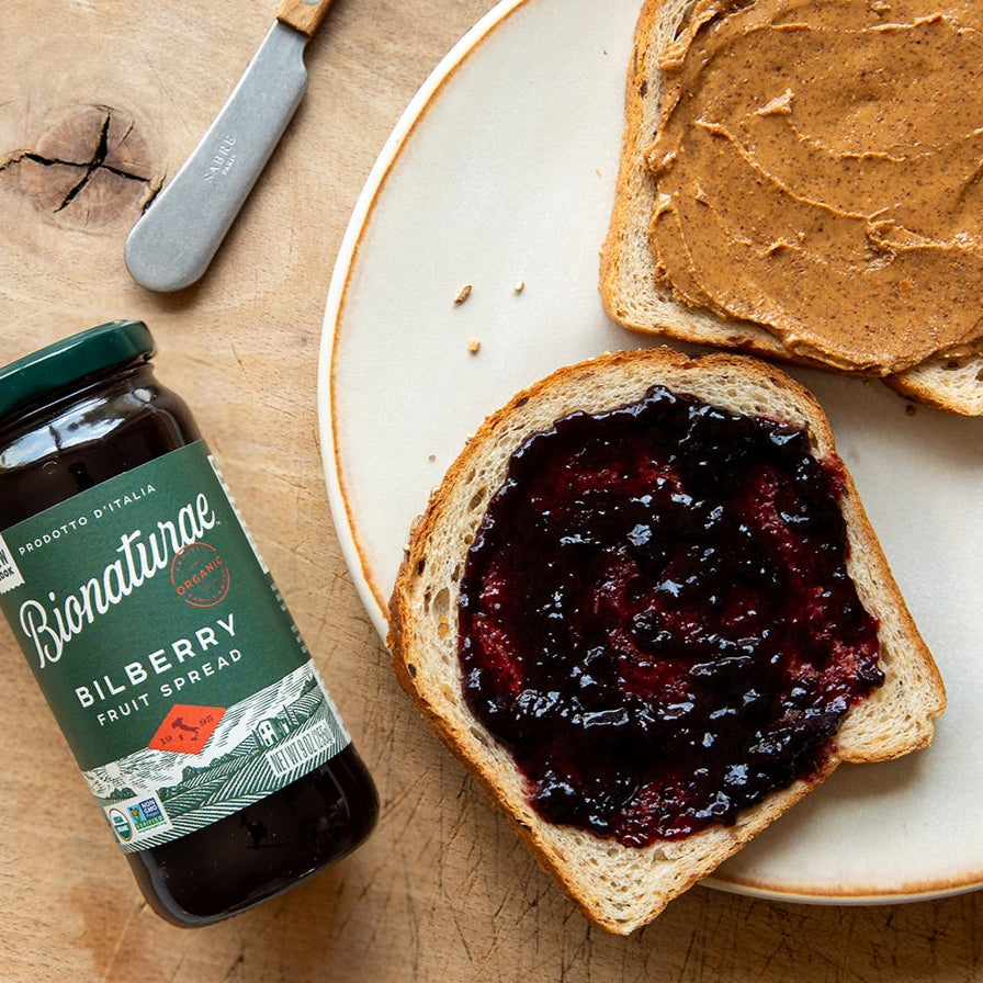 Jar of Bionaturae organic bilberry fruit spread beside a slice of bread topped with bilberry spread on a wooden table